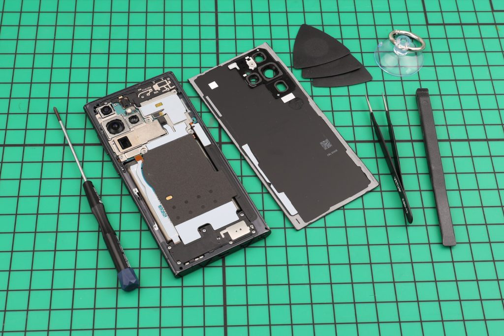 Samsung phone opened on a green cutting mat, back cover lifted off beside tweezers and picks