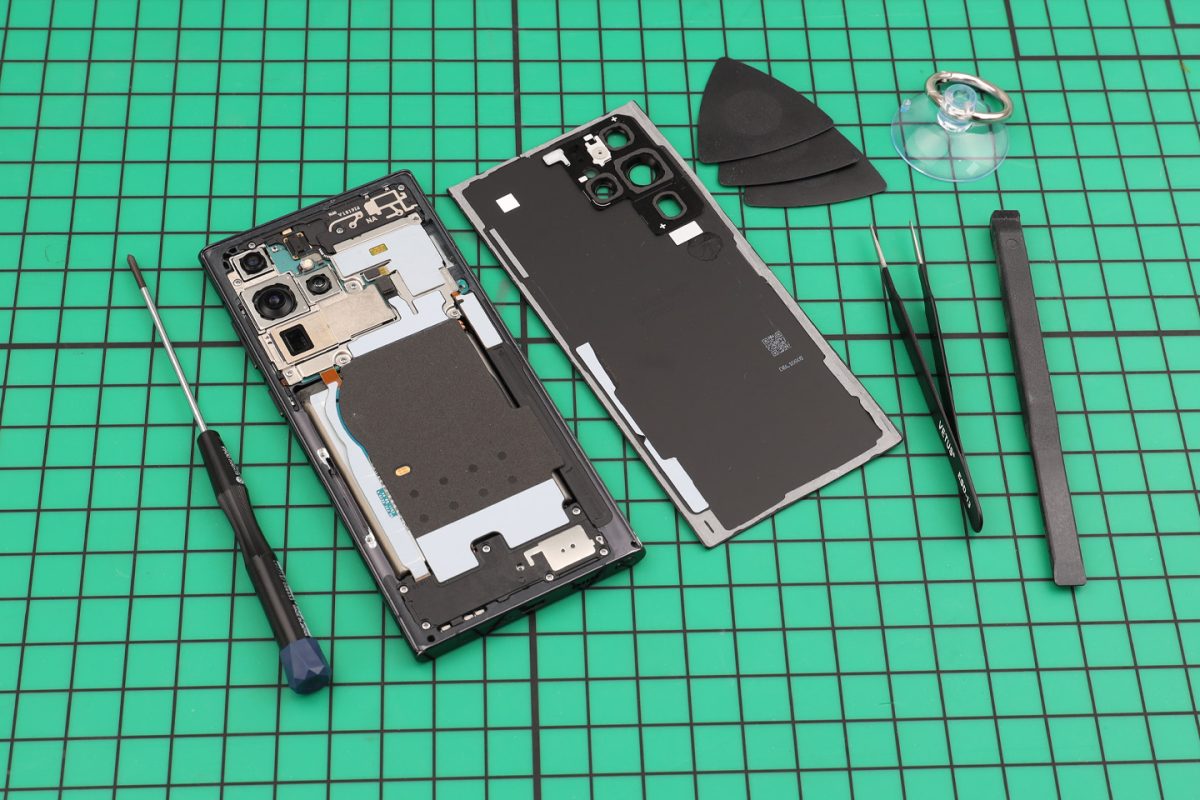 Samsung phone opened on a green cutting mat, back cover lifted off beside tweezers and picks