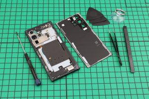 Samsung phone opened on a green cutting mat, back cover lifted off beside tweezers and picks