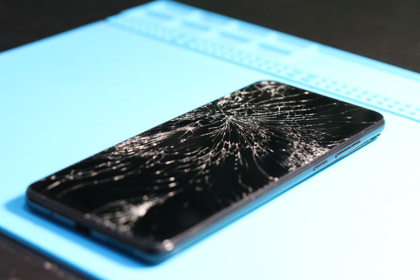 Black smartphone with a spiderweb cracked display resting on a blue silicone repair mat