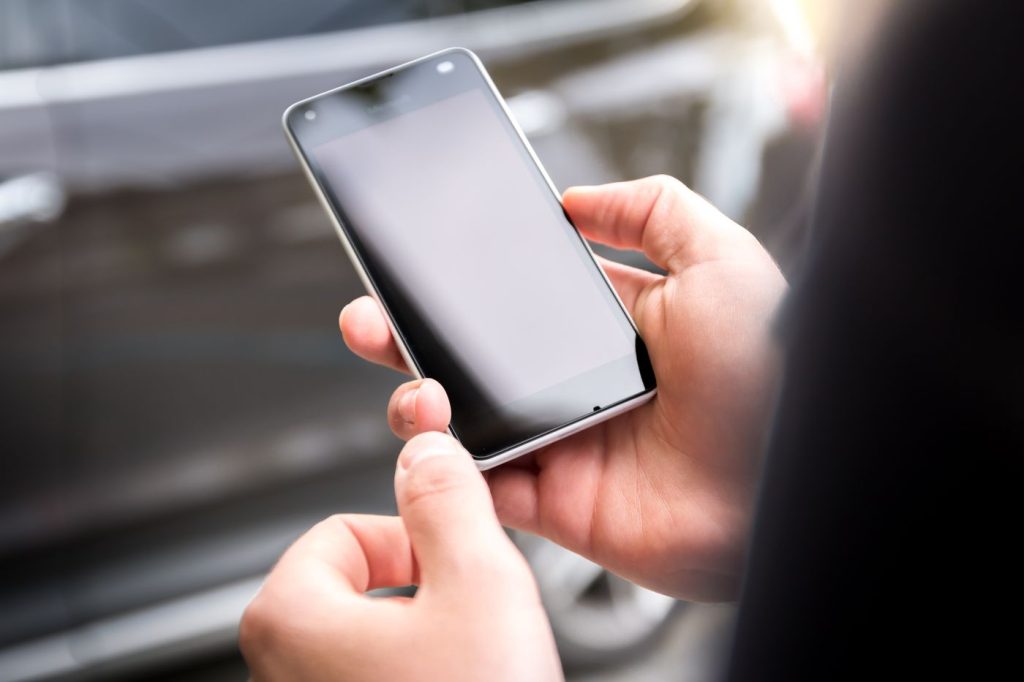 Two hands holding a dark smartphone with a blank, unlit screen outdoors in daylight